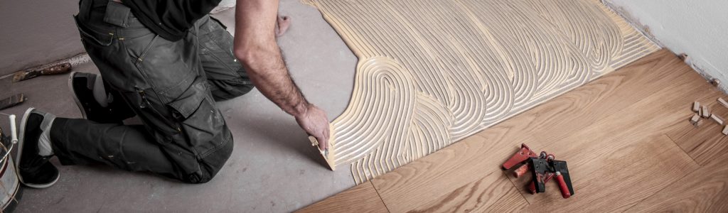 Handwerker verlegt Parkettboden und streicht den Estrich mit Kleber ein und schlägt die Dielen mit dem Hammer und dem Schlagklotz zusammen Handwerker bestreicht den Estrich mit Kleber bevor er den Parkettboden verlegt